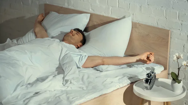 Пробуджений чоловік посміхається, розтягуючись в ліжку біля будильника на тумбочці — стокове фото