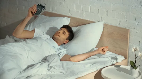 Hombre sonriente mirando el reloj despertador vintage mientras está acostado en la cama por la mañana - foto de stock