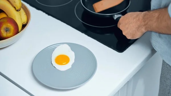 Cropped view of man with frying pan and spatula near fried egg on plate and fruits in bowl — Stock Photo