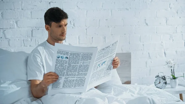 Hombre leyendo noticias económicas en la cama cerca del despertador y orquídea en maceta en la mesita de noche - foto de stock