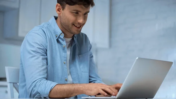 Freelance sorridente in camicia blu che scrive su computer portatile in cucina — Foto stock