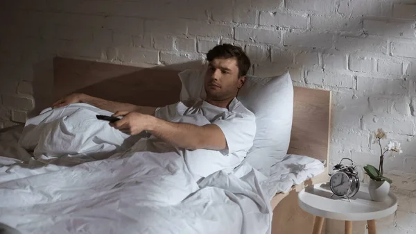 Homem clicando canais enquanto assiste tv na cama perto de despertador e orquídea na mesa de cabeceira — Fotografia de Stock