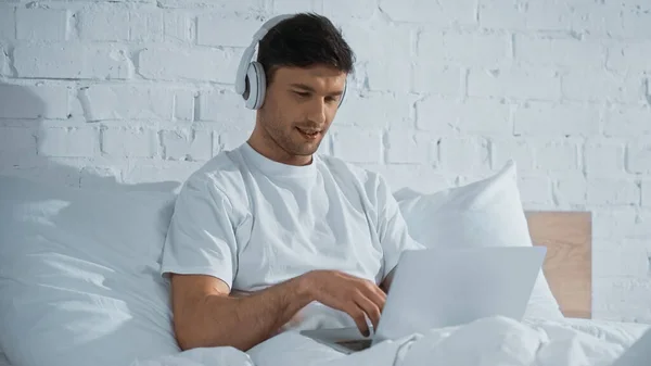 Hombre en auriculares escribiendo en el ordenador portátil mientras está sentado en la cama - foto de stock