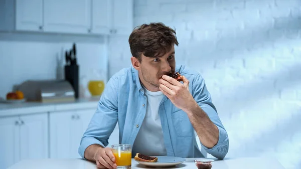Hombre comiendo tostadas con confitura cerca de vaso de jugo de naranja en la cocina - foto de stock