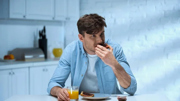 Uomo mangiare pane tostato con confiture vicino a vetro di succo d'arancia in cucina — Foto stock