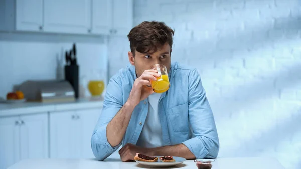 Человек в голубой рубашке пьет апельсиновый сок рядом с тостами со сладким джемом на кухне — стоковое фото
