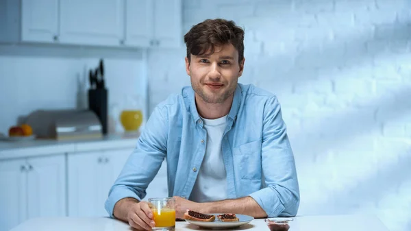 Homme heureux regardant la caméra tout en étant assis près de savoureux petit déjeuner dans la cuisine — Photo de stock