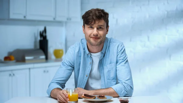 Felice uomo guardando la fotocamera mentre seduto vicino gustosa colazione in cucina — Foto stock