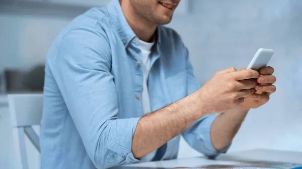 Обрезанный вид улыбающегося мужчины в синей рубашке, который пишет сообщения по сотовому телефону на кухне — стоковое фото