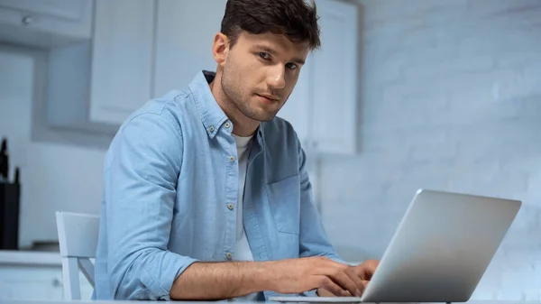 Freelance en chemise bleue regardant la caméra tout en utilisant un ordinateur portable dans la cuisine — Photo de stock