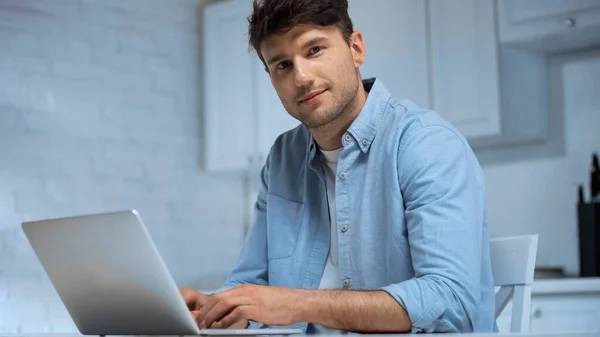 Positiver Mann im blauen Hemd blickt in die Kamera, während er in der Küche auf dem Laptop tippt — Stockfoto