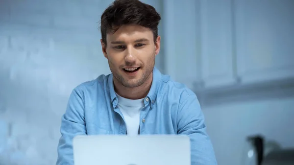 Uomo sorridente in camicia blu che lavora su laptop in cucina — Foto stock