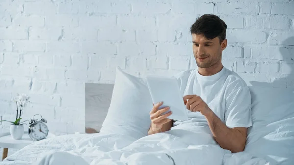 Uomo sorridente utilizzando tablet digitale mentre seduto a letto vicino sveglia sul comodino — Foto stock