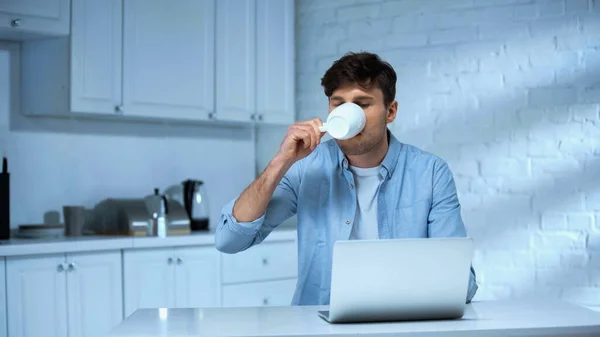 Freelancer em camisa azul beber café perto de laptop na cozinha — Fotografia de Stock