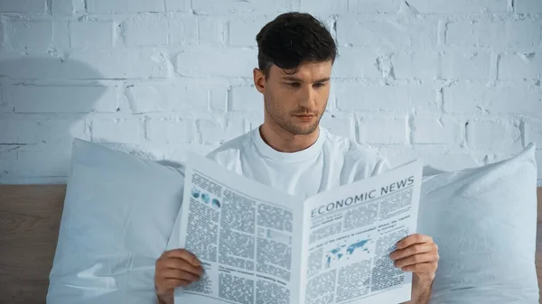 Hombre leyendo noticias económicas mientras está sentado en la cama por la mañana - foto de stock