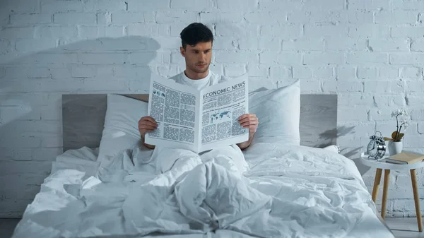 Homem lendo jornal enquanto sentado na cama sob cobertor branco — Fotografia de Stock