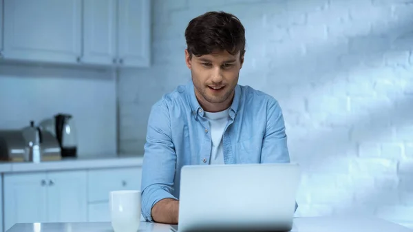 Man in blue shirt smiling while working on laptop in kitchen — Stock Photo