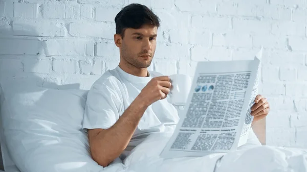 Homem com copo leitura jornal da manhã enquanto sentado na cama de manhã — Fotografia de Stock