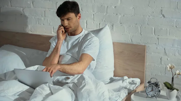 Homem apontando para laptop enquanto fala no smartphone na cama — Fotografia de Stock