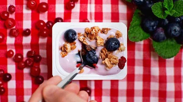 Cropped view of man holding spoon near yogurt with granola and berries in plastic container — Stock Photo