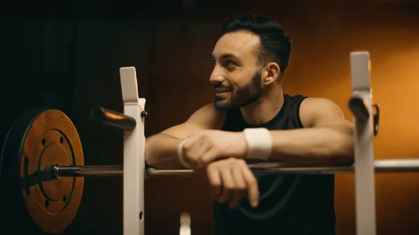 Smiling bearded sportsman standing near barbell on stand on dark background — Stock Photo
