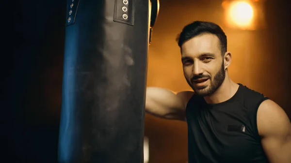 Улыбающийся спортсмен смотрит в камеру рядом с боксёрской грушей на тёмном фоне — стоковое фото