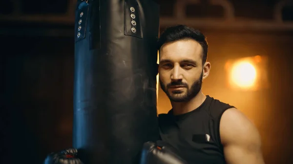 Bearded sportsman in gloves looking at camera near punch bag on dark background — Stock Photo