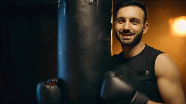 Deportista positivo en guantes de boxeo mirando a la cámara cerca del saco de boxeo sobre fondo oscuro - foto de stock