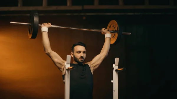 Muscular sportsman training with barbell near stand on dark background — Stock Photo