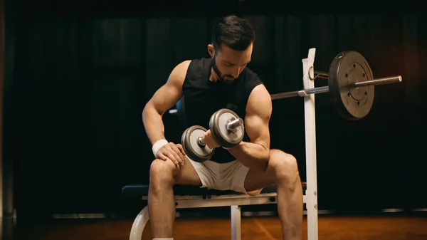 Muscular sportsman training with dumbbell on flat bench on dark background — Stock Photo