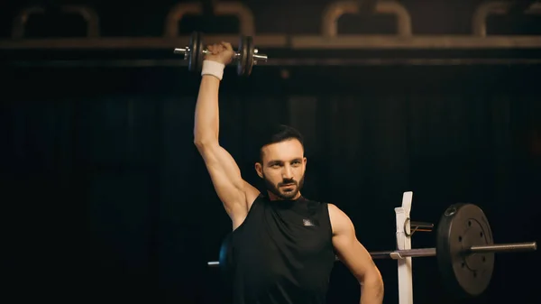 Athletic sportsman working out with dumbbell near barbell on dark background — Stock Photo