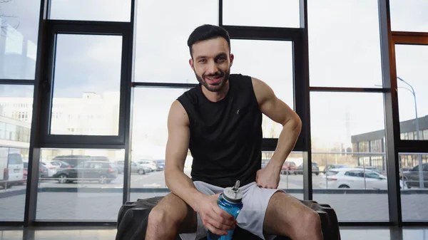 Sportif souriant tenant une bouteille de sport assis sur un pneu dans une salle de gym — Photo de stock