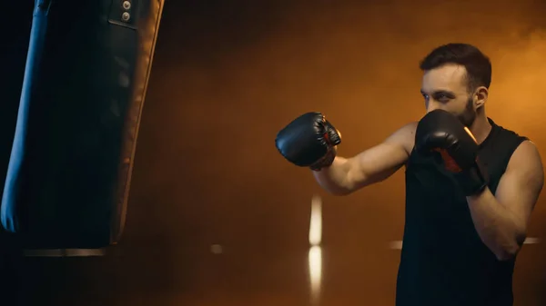 Sportler in Boxhandschuhen beim Training mit Boxsack auf dunklem Hintergrund — Stockfoto