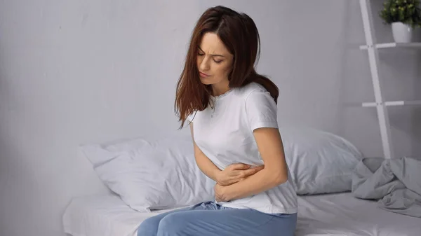 Woman sitting on bed and hugging belly while feeling abdominal pain — Stock Photo