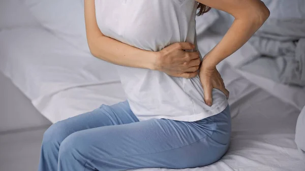 Partial view of woman in pajamas touching painful loin while sitting on bed — Stock Photo