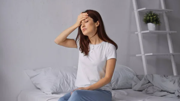 Brünette Frau mit Kopfschmerzen, die Stirn berührt, während sie mit geschlossenen Augen auf dem Bett sitzt — Stockfoto