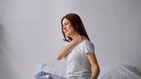 Brunette woman with closed eyes sitting on bed and touching painful neck — Stock Photo