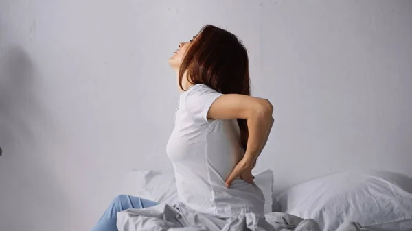 Visão lateral da mulher que sofre de dor nas costas enquanto está sentada na cama — Fotografia de Stock