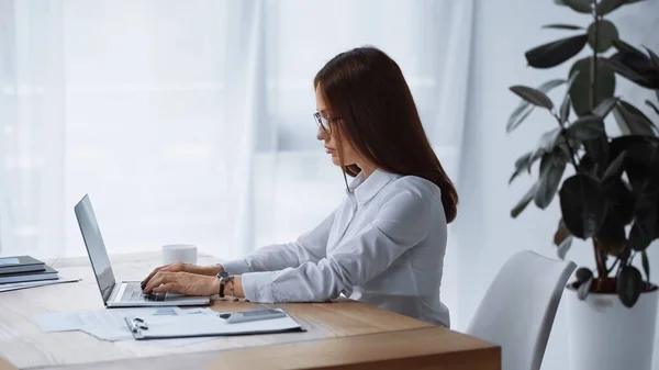 Seitenansicht einer brünetten Frau, die im Büro arbeitet und auf dem Laptop tippt — Stockfoto