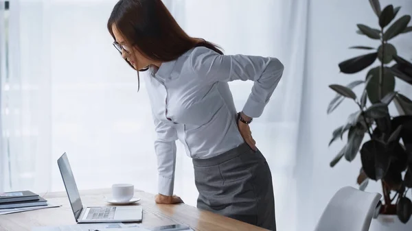 Brunette woman touching painful back while standing at work desk — Stock Photo