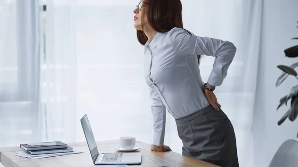 Brunette woman standing at work desk and touching painful loin — Stock Photo