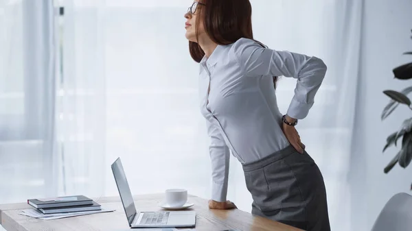 Morena mulher de pé na mesa de trabalho e tocando doloroso lombo — Fotografia de Stock