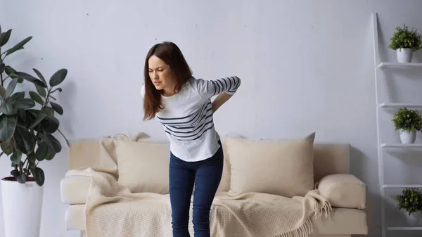 Brünette Frau mit geschlossenen Augen, die neben Sofa steht und schmerzenden Rücken berührt — Stockfoto