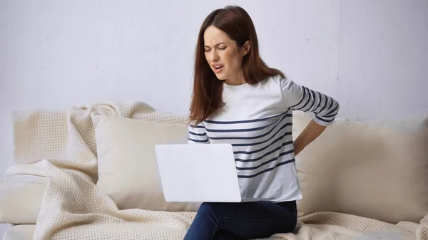 Незадоволена жінка сидить на дивані з ноутбуком і торкається болючої пов'язки — стокове фото