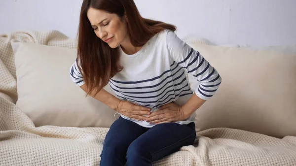 Woman with closed eyes suffering from stomach pain while sitting on sofa — Stock Photo