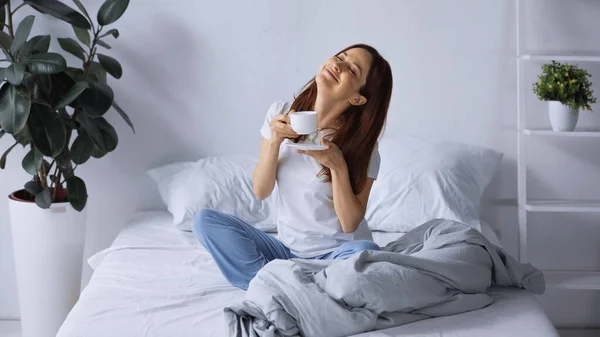 Cheerful woman in pajamas sitting on bed with closed eyes and holding cup of coffee — Stock Photo