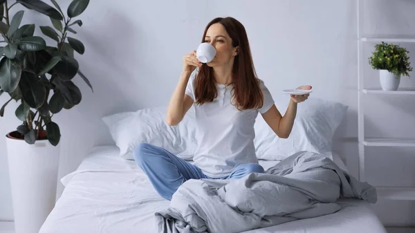 Brünette Frau im Pyjama trinkt Morgenkaffee, während sie auf dem Bett sitzt — Stockfoto