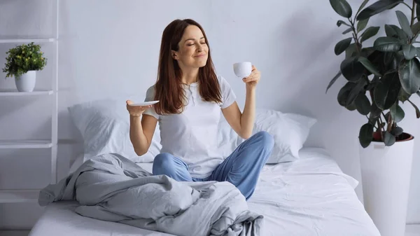 Mulher sorridente em pijama segurando xícara de café enquanto sentado na cama com os olhos fechados — Fotografia de Stock