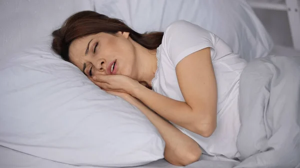 Mujer frunciendo el ceño acostada en la cama con los ojos cerrados y sufriendo de dolor de muelas - foto de stock