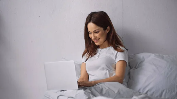 Brunette woman smiling while using laptop on bed at home — Stock Photo